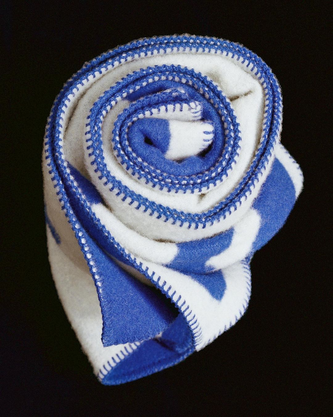 Close-up of a rolled, soft white blanket with blue stripes and blue overcast stitching edges, set against a dark backdrop.