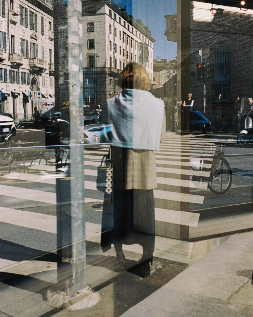 Woman in coat stands on a city crosswalk, reflected in a window with street scene, showcasing urban life and architectural backdrop.