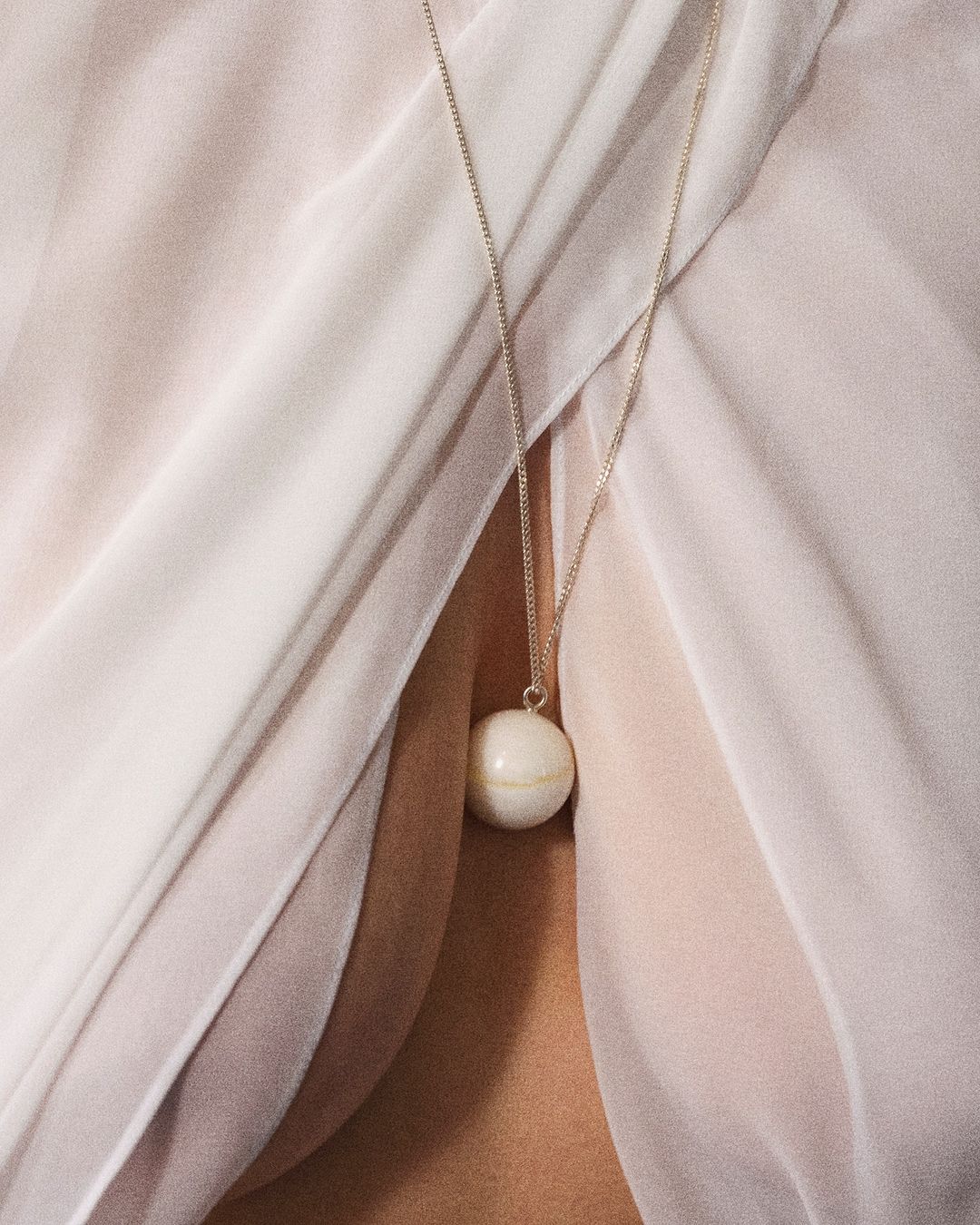 Close-up of a woman wearing a white ball pendant necklace on a silver chain, set against a pale pink draped fabric background.