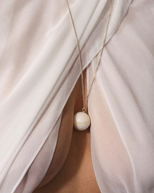 Close-up of a woman wearing a white ball pendant necklace on a silver chain, set against a pale pink draped fabric background.