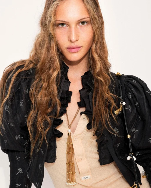 Portrait of a young woman with long, wavy auburn hair wearing a black floral jacket and a beige button-down shirt with a gold necklace.