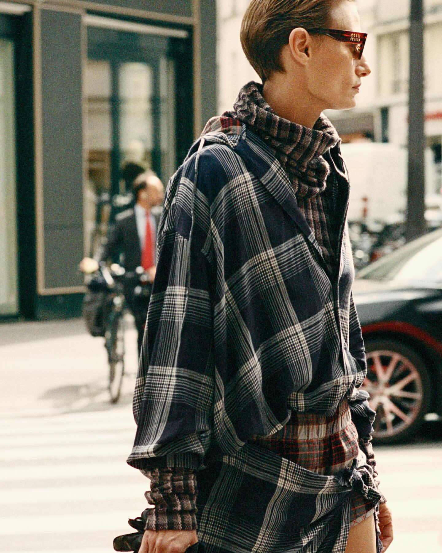 Woman in oversized plaid shirt and sunglasses standing on a city street with car and cyclist in background, fashion editorial shot.