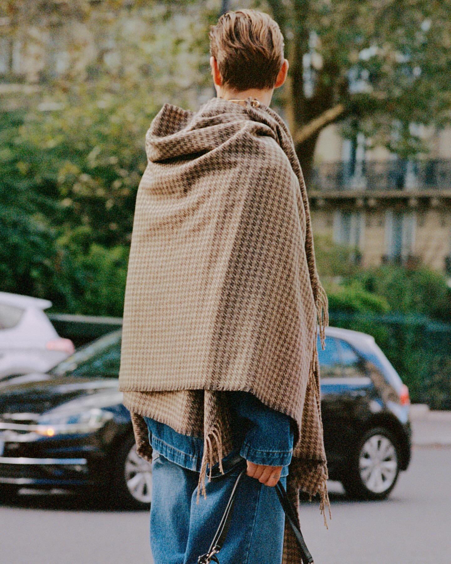 Back view of a slender figure wearing oversized denim and a patterned poncho, standing on a city street with cars and buildings.