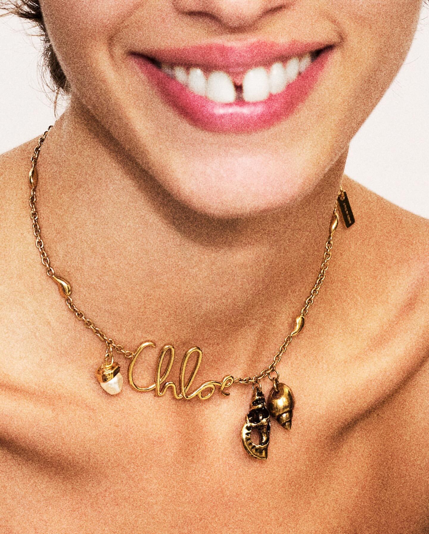 Close-up shot of a woman smiling, showcasing a gold 'Chloe' necklace with shell charms against a neutral background. Beauty and fashion.