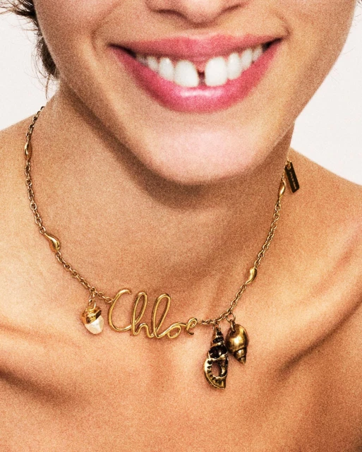 Close-up shot of a woman smiling, showcasing a gold 'Chloe' necklace with shell charms against a neutral background. Beauty and fashion.