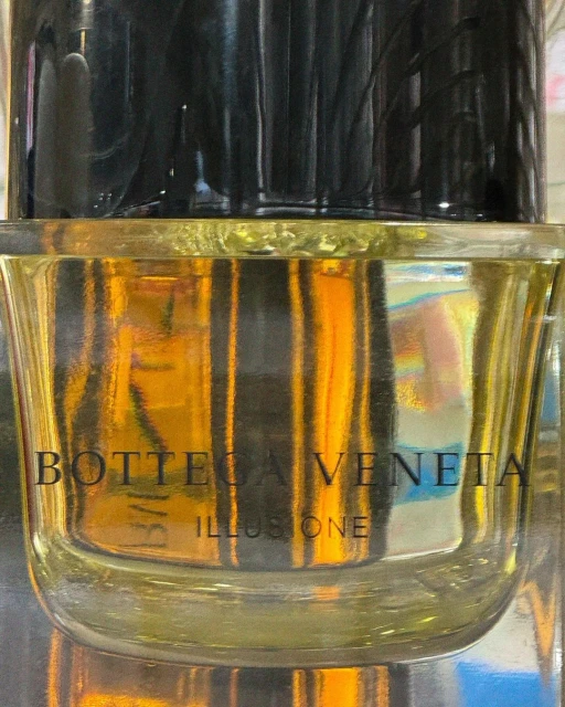 Close-up shot of Bottega Veneta Illusione perfume bottle with a clear glass container and a black stopper, showcasing the brand's logo.