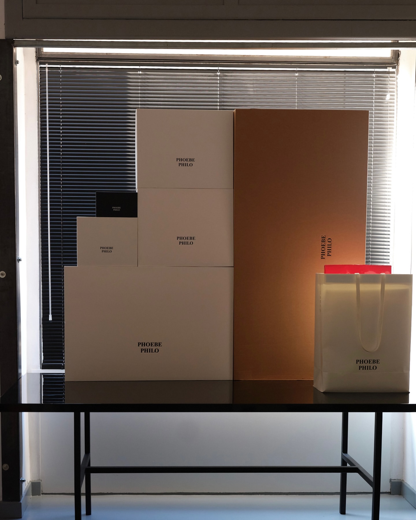 Assortment of branded boxes and a shopping bag, all featuring the 'Phoebe Philo' logo, arranged on a black table against a window backdrop.