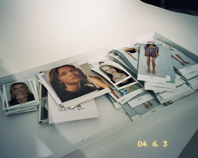 Model Polaroids are arranged in a tray; several young adult females are featured in the photos. The polaroids appear to be casting photos.