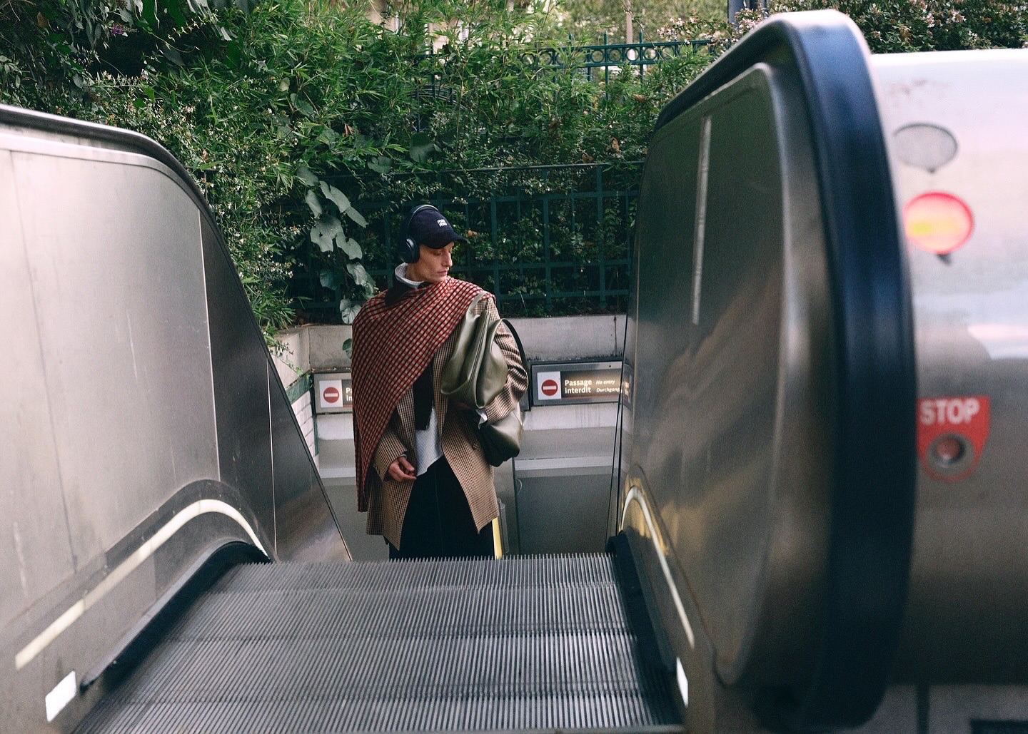 Young adult on escalator wearing headphones, draped in red patterned shawl, brown jacket, black pants, walking toward subway entrance.