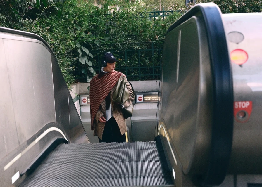 Young adult on escalator wearing headphones, draped in red patterned shawl, brown jacket, black pants, walking toward subway entrance.