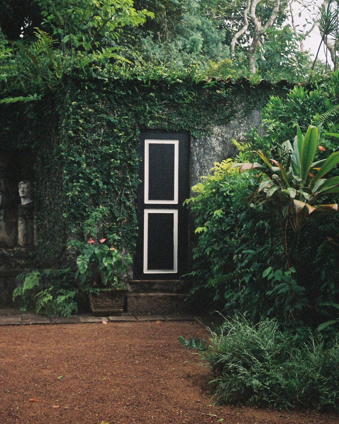Exterior of stone building covered in ivy with black door and red-brown gravel ground.