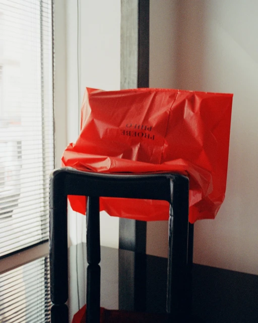 Red plastic bag sits on a black stool, illuminated by window light, in an indoor setting with monochrome accents. Modern shot.