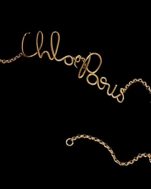 Close-up of a gold necklace spelling out 'Chloe Paris' in cursive script against a stark black background, showcasing jewelry.