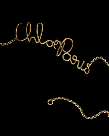 Close-up of a gold necklace spelling out 'Chloe Paris' in cursive script against a stark black background, showcasing jewelry.