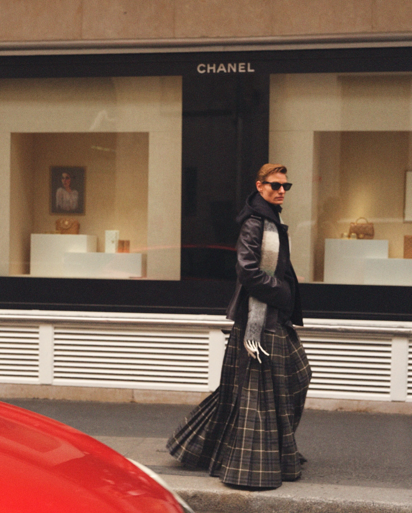 A fashionable man in a kilt walks by a Chanel storefront, wearing sunglasses, a leather jacket, and a scarf in an urban setting.