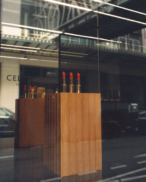 Three red lipsticks displayed on a wooden stand in a storefront window, reflecting the city street and buildings outside.