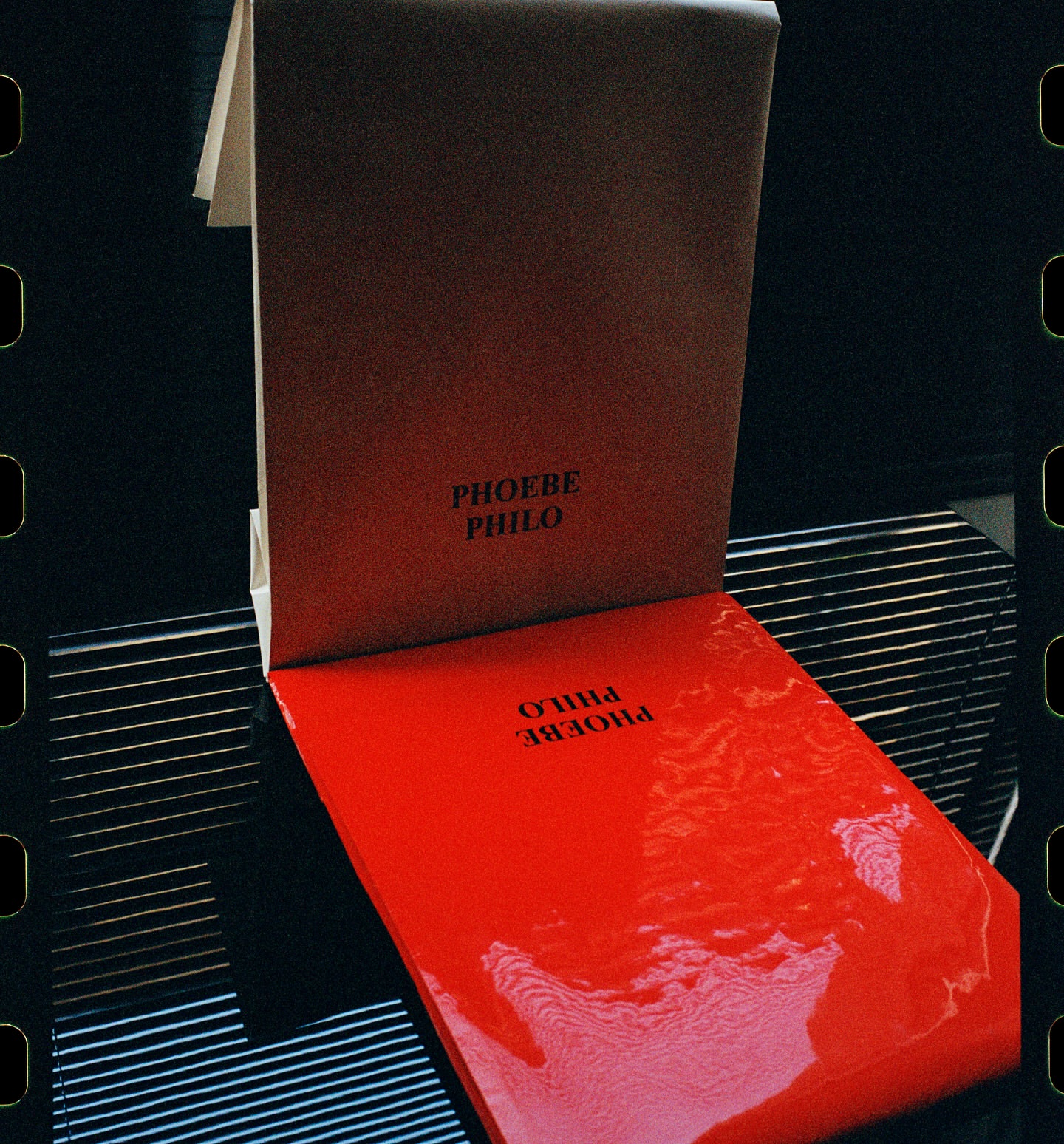 Two vibrant Phoebe Philo books, one red and one brown, stacked on a reflective surface with subtle lighting and a film border.