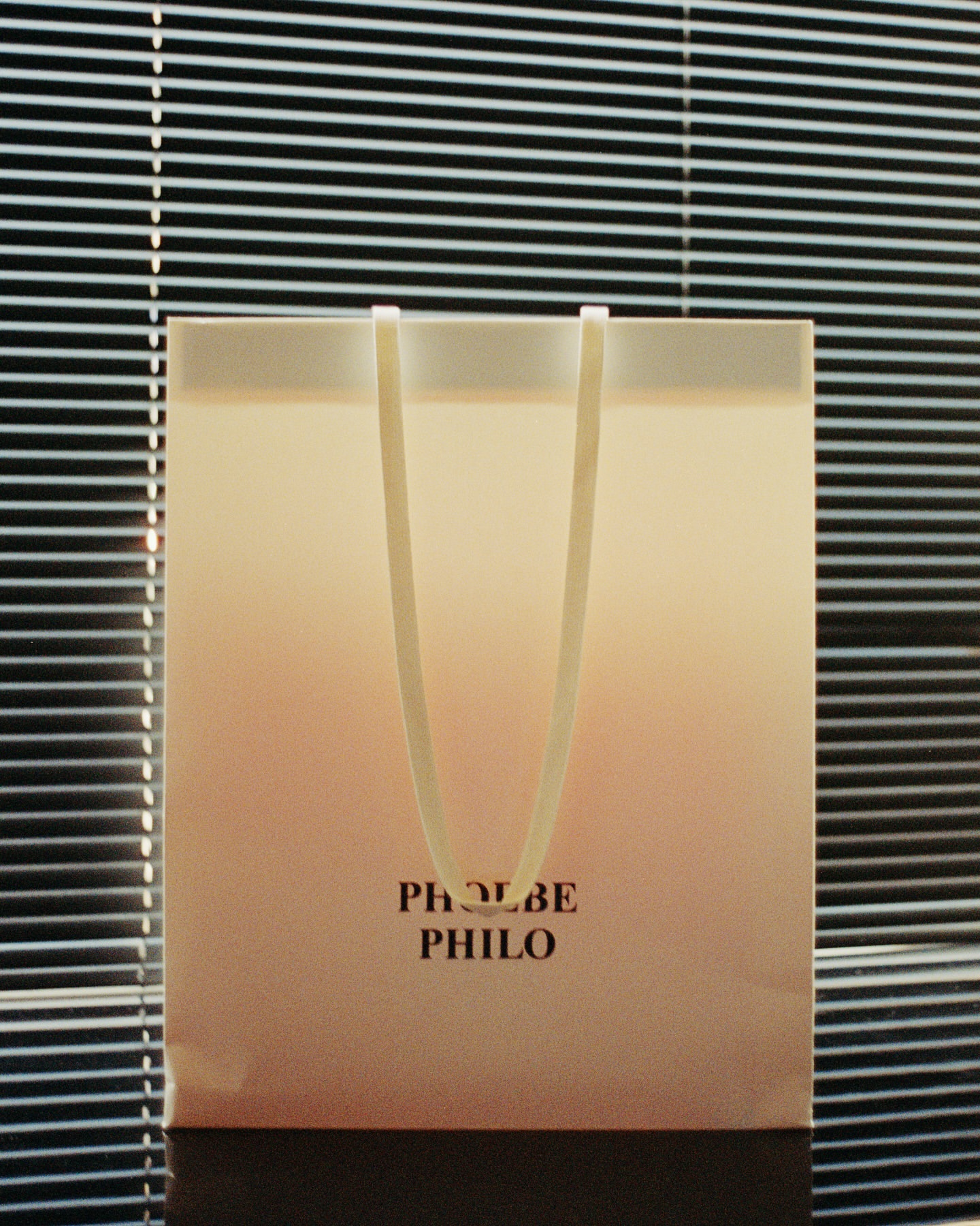 Close-up of a shopping bag with the text 'PHOEBE PHILO' on it, set against a backdrop of closed blinds, soft lighting.