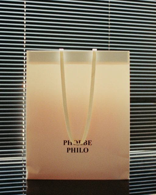 Close-up of a shopping bag with the text 'PHOEBE PHILO' on it, set against a backdrop of closed blinds, soft lighting.
