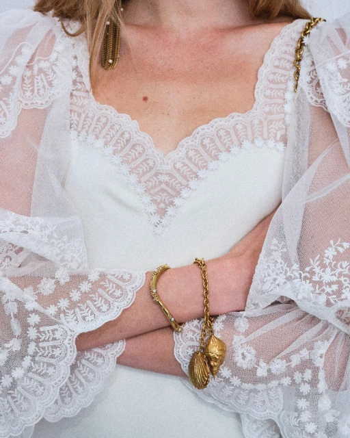 Close-up shot of a woman's torso wearing a white lace dress, gold chain earrings, and a shell bracelet with her arms crossed.