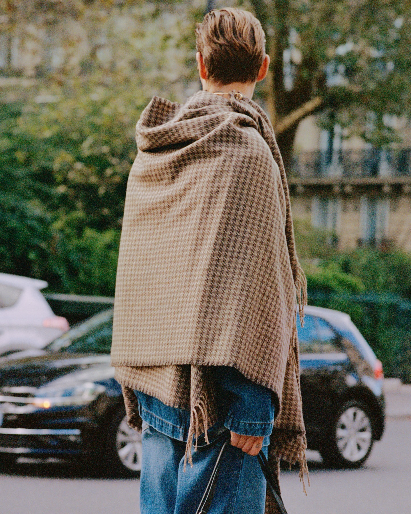 Rear view of a person wearing a brown houndstooth blanket scarf and blue jeans against a city street backdrop.