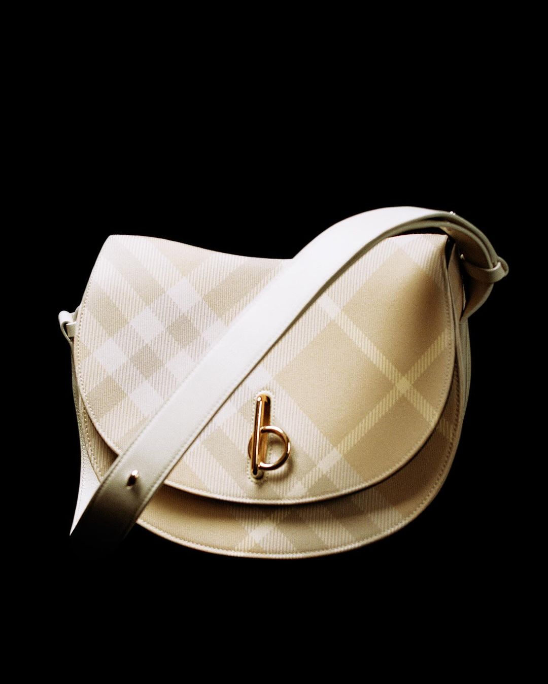 Beige saddle bag with a plaid pattern and white shoulder strap on a black background, perfect for fashion-forward individuals.