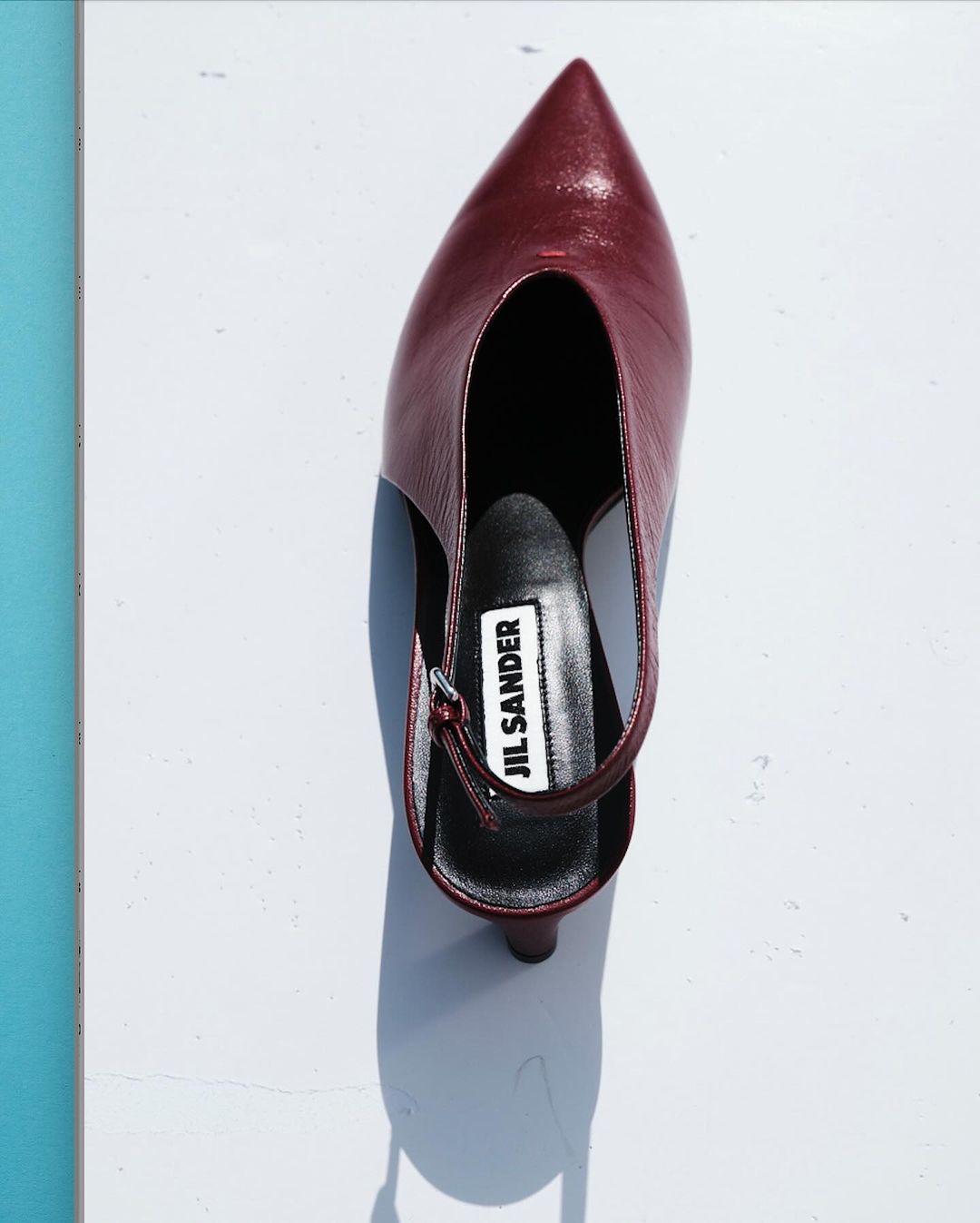 Burgundy Jil Sander slingback heel on a white surface, angled overhead shot. Formal shoe with a pointy toe and slingback strap.