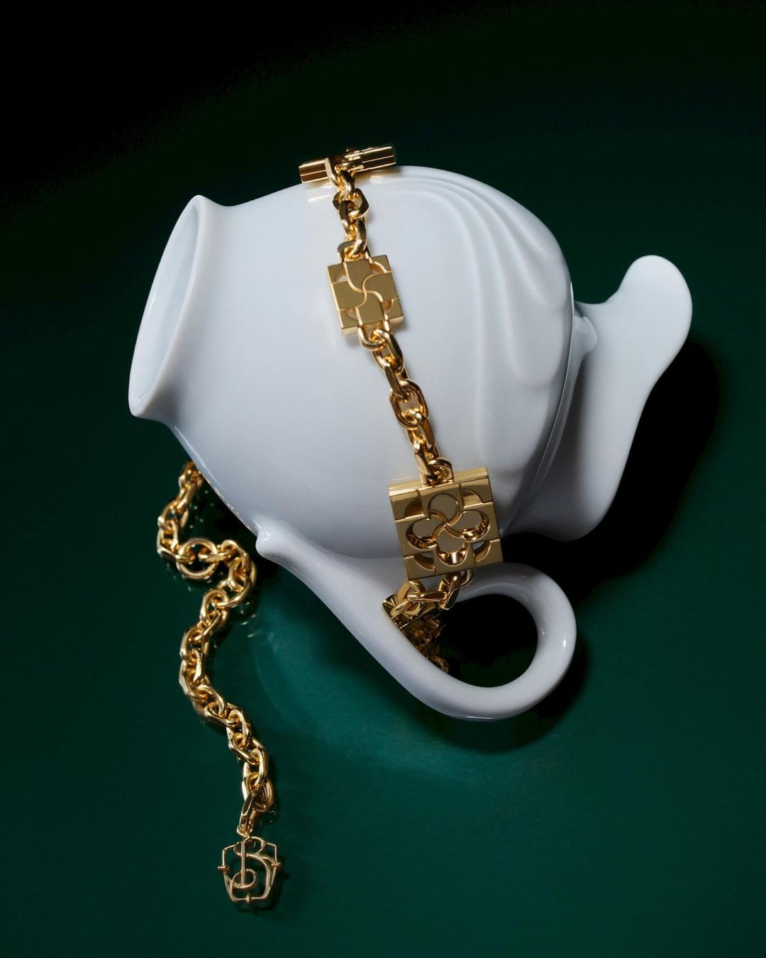 Gold chain necklace draped over a white ceramic teapot against a dark green backdrop in a high-angle studio shot.