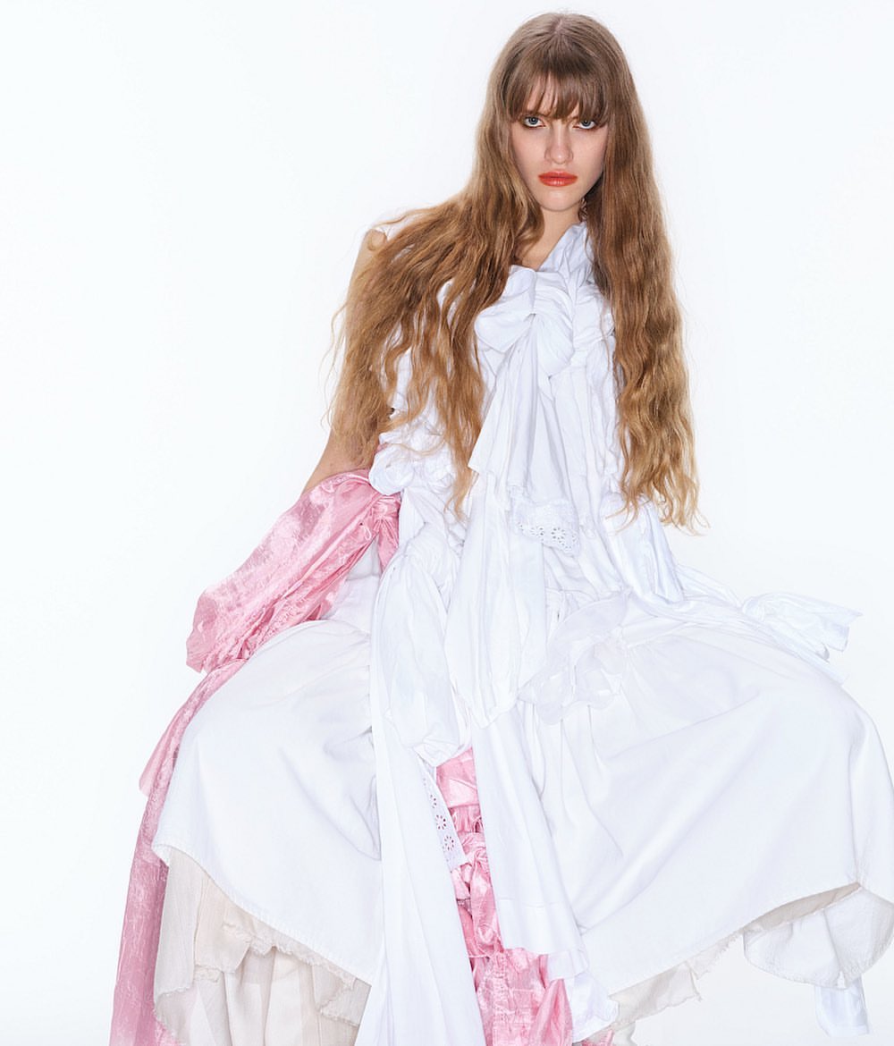 A woman with long brown hair sits surrounded by layered white and pink fabric against a white background in a fashion editorial shot.