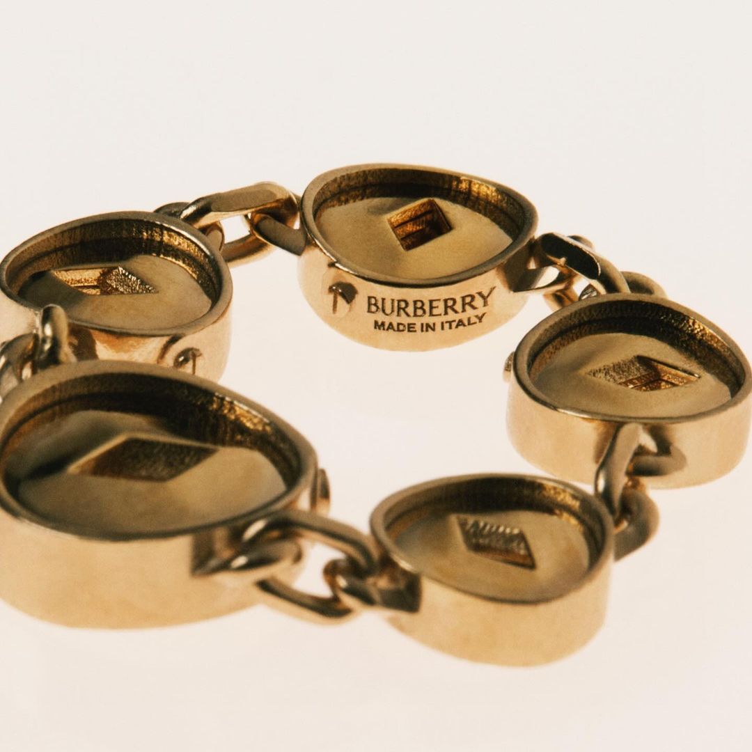 Close-up of a gold Burberry chain bracelet featuring circular links with engraved Burberry branding on a neutral background. Fashion jewelry.
