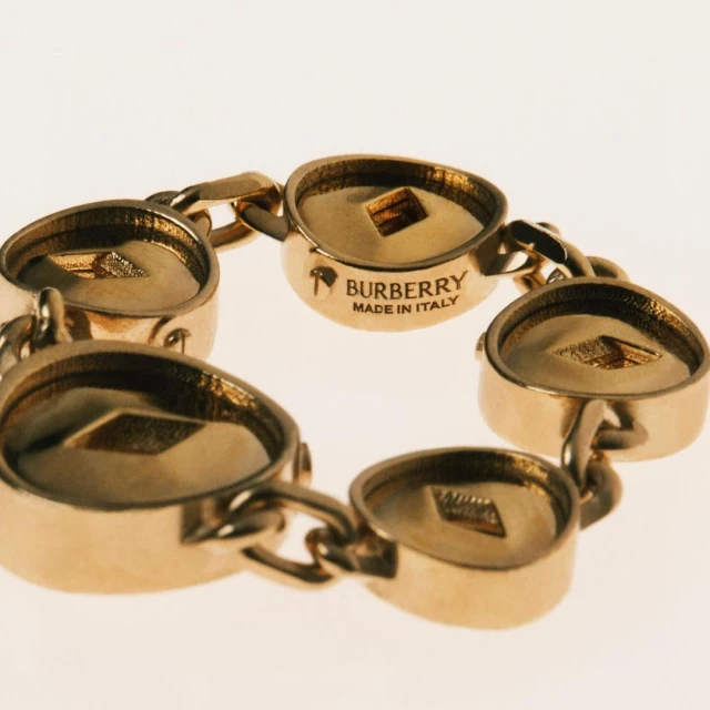 Close-up of a gold Burberry chain bracelet featuring circular links with engraved Burberry branding on a neutral background. Fashion jewelry.
