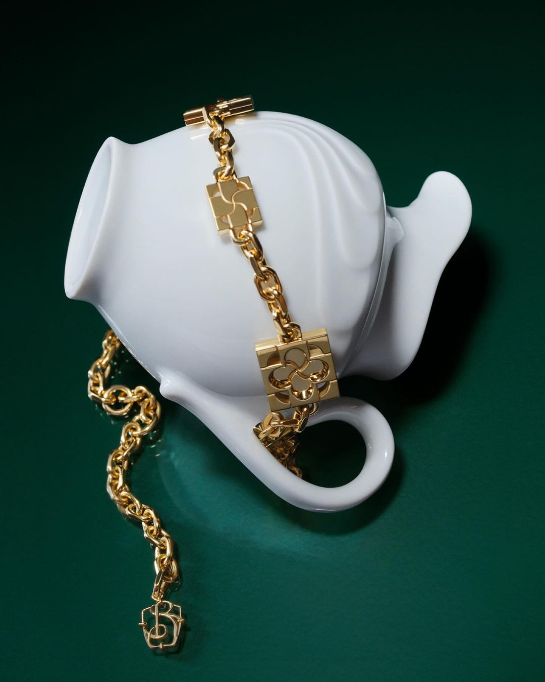 Elegant gold chain necklace draped over a white ceramic teapot against a deep green backdrop in a softly lit studio setting.