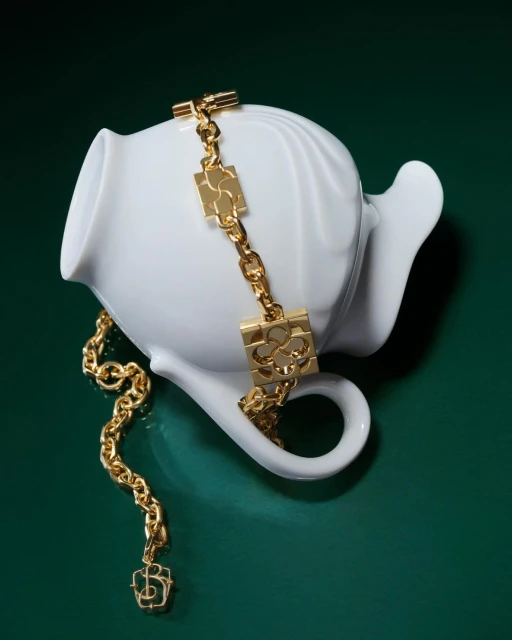 Elegant gold chain necklace draped over a white ceramic teapot against a deep green backdrop in a softly lit studio setting.