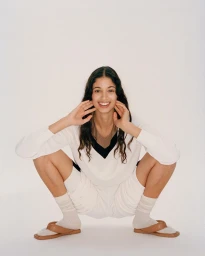 Smiling woman in white v-neck sweater and shorts squatting against a plain white background