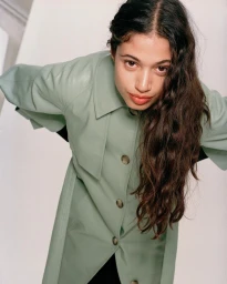 Portrait of a woman wearing a pale green button-up coat looking down towards the camera against a neutral studio background.