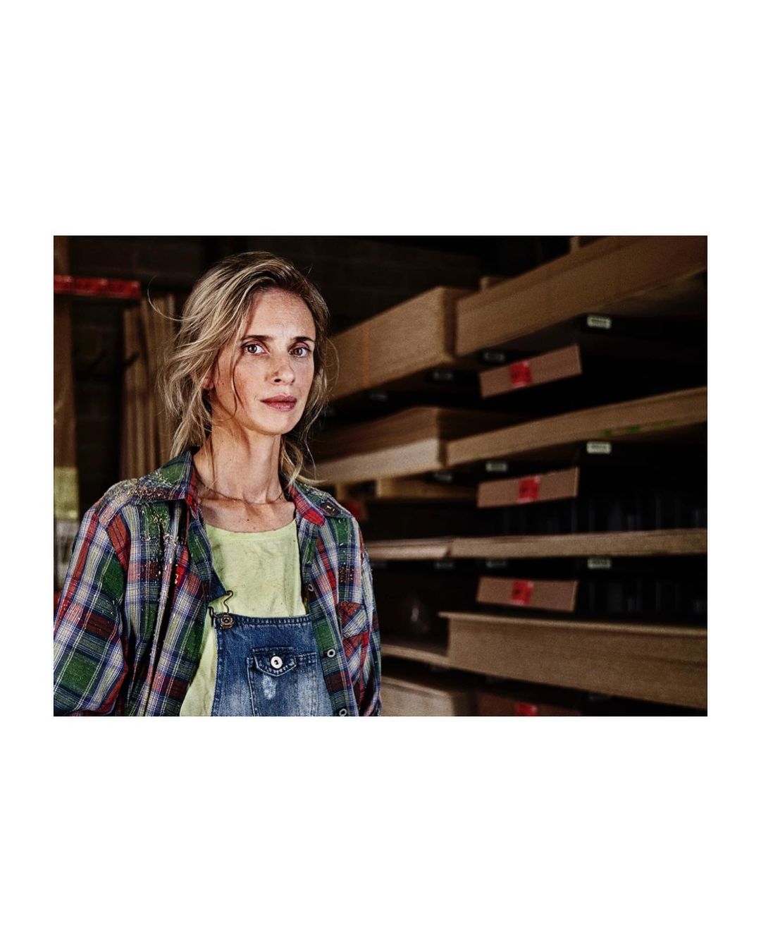 Portrait of a woman in overalls and a flannel shirt standing in what appears to be a lumber or woodworking warehouse setting.