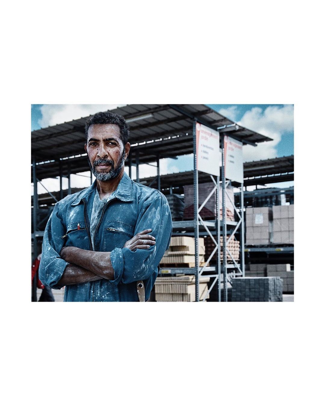 Adult male worker in blue coveralls stands with arms crossed in a warehouse setting, surrounded by shelves of construction materials.