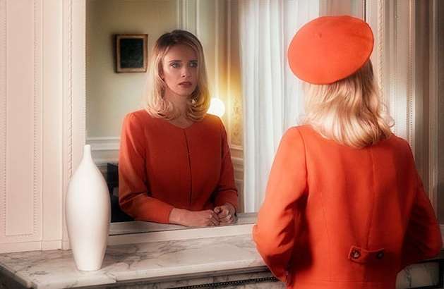 Woman in an orange dress and beret looking in a mirror in an indoor setting with a white vase on a marble surface.
