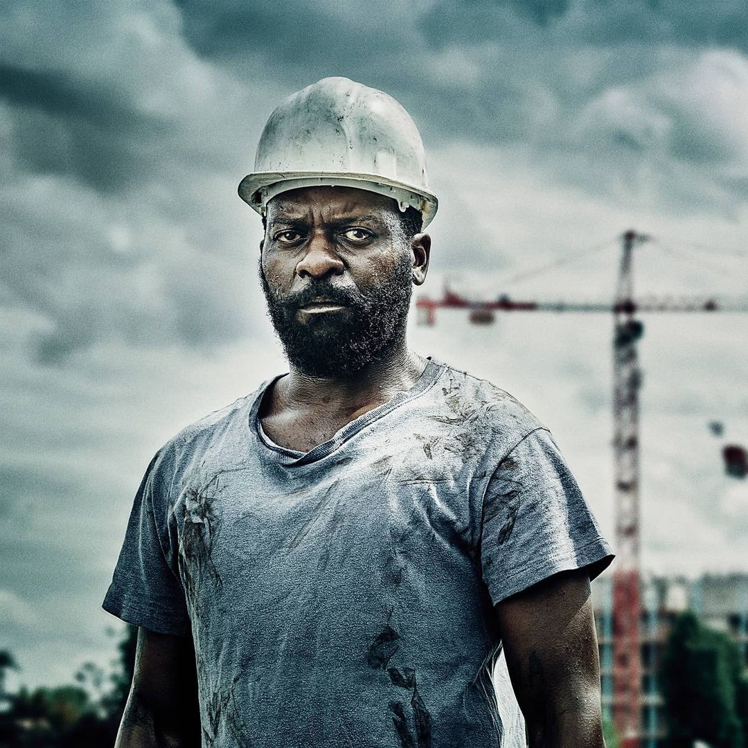 Portrait of a serious African-American construction worker in a white hardhat and grey t-shirt, with a crane and cloudy sky backdrop.