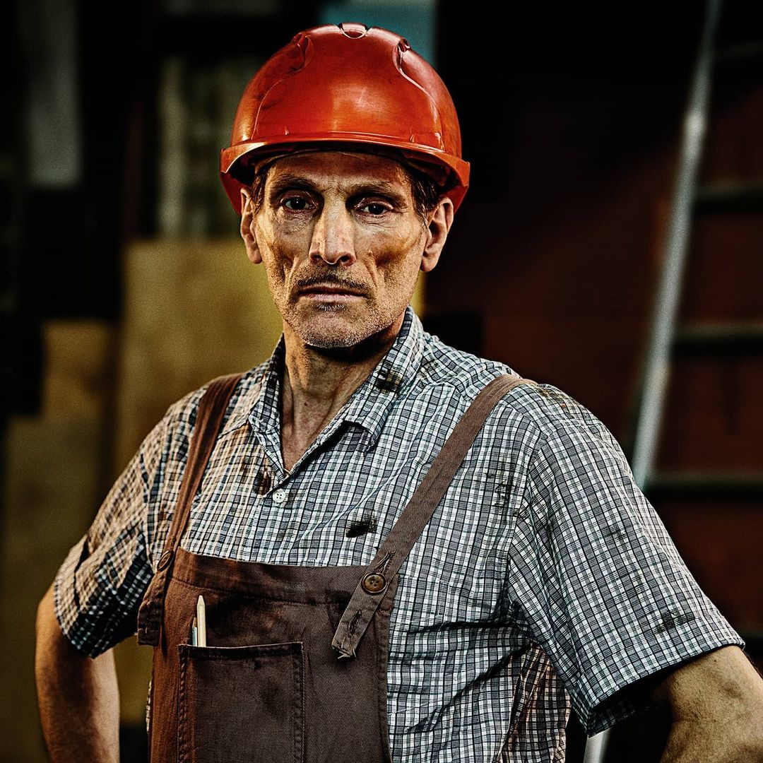 Portrait of a construction worker wearing a red hard hat and brown overalls with a pen in his pocket standing indoors.
