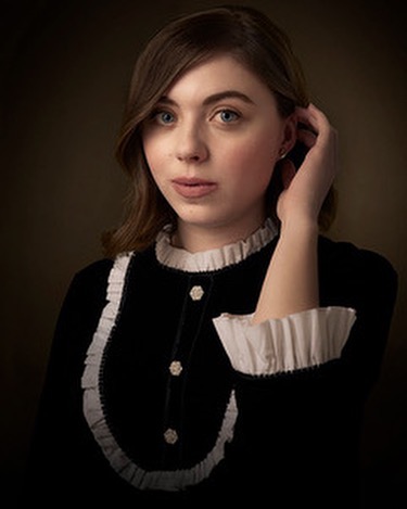 Portrait of a fair-skinned young woman with wavy brown hair, wearing a black dress with white ruffled trim, posing in a studio setting.