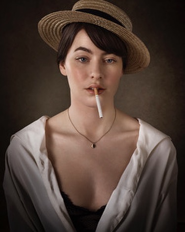 Portrait of a woman wearing a straw hat, holding a cigarette in her mouth, against a dark backdrop with a white collared shirt.