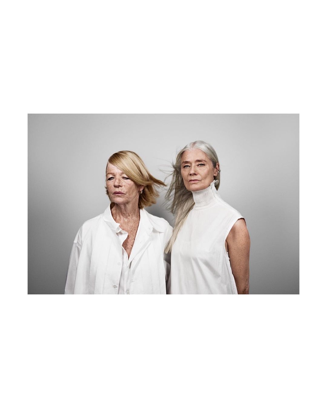 Two mature women pose in white against a gray background. One has blonde hair, the other gray. Fashion and beauty portrait, studio shot.