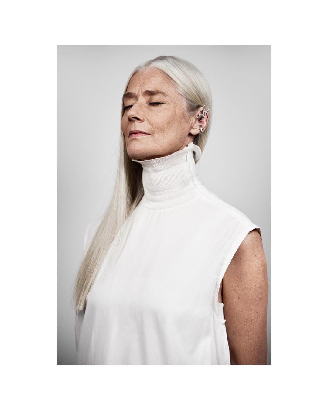 Portrait of an older woman with gray hair and closed eyes, wearing a white top against a light gray background, highlighting natural beauty.