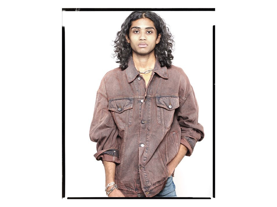 Young adult poses indoors wearing a vintage denim jacket against a white backdrop, showcasing fashion and casual style.