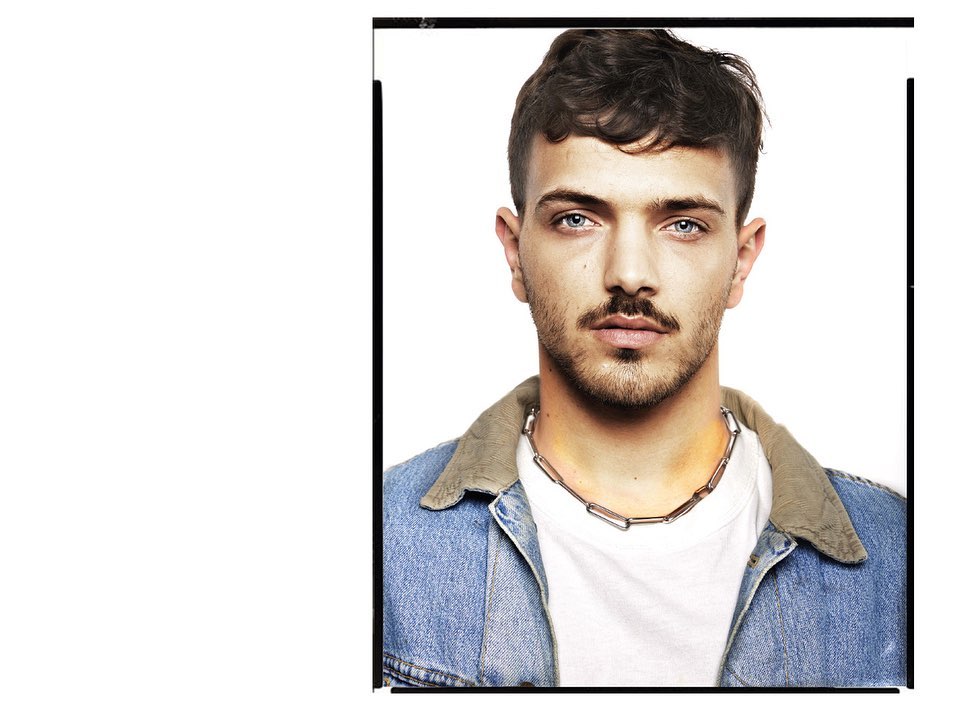 Portrait of a young adult male with blue eyes wearing a denim jacket and chain necklace against a white background.
