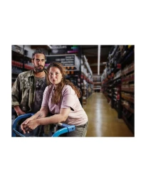A man and a woman posing inside a large hardware store, with the woman leaning on a blue shopping cart handle.