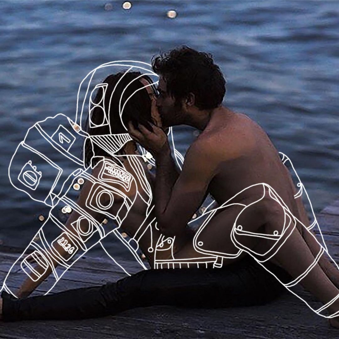 Couple sitting together on a wooden platform near the water at dusk, one outlined in glowing white as an astronaut. A romantic artistic moment.