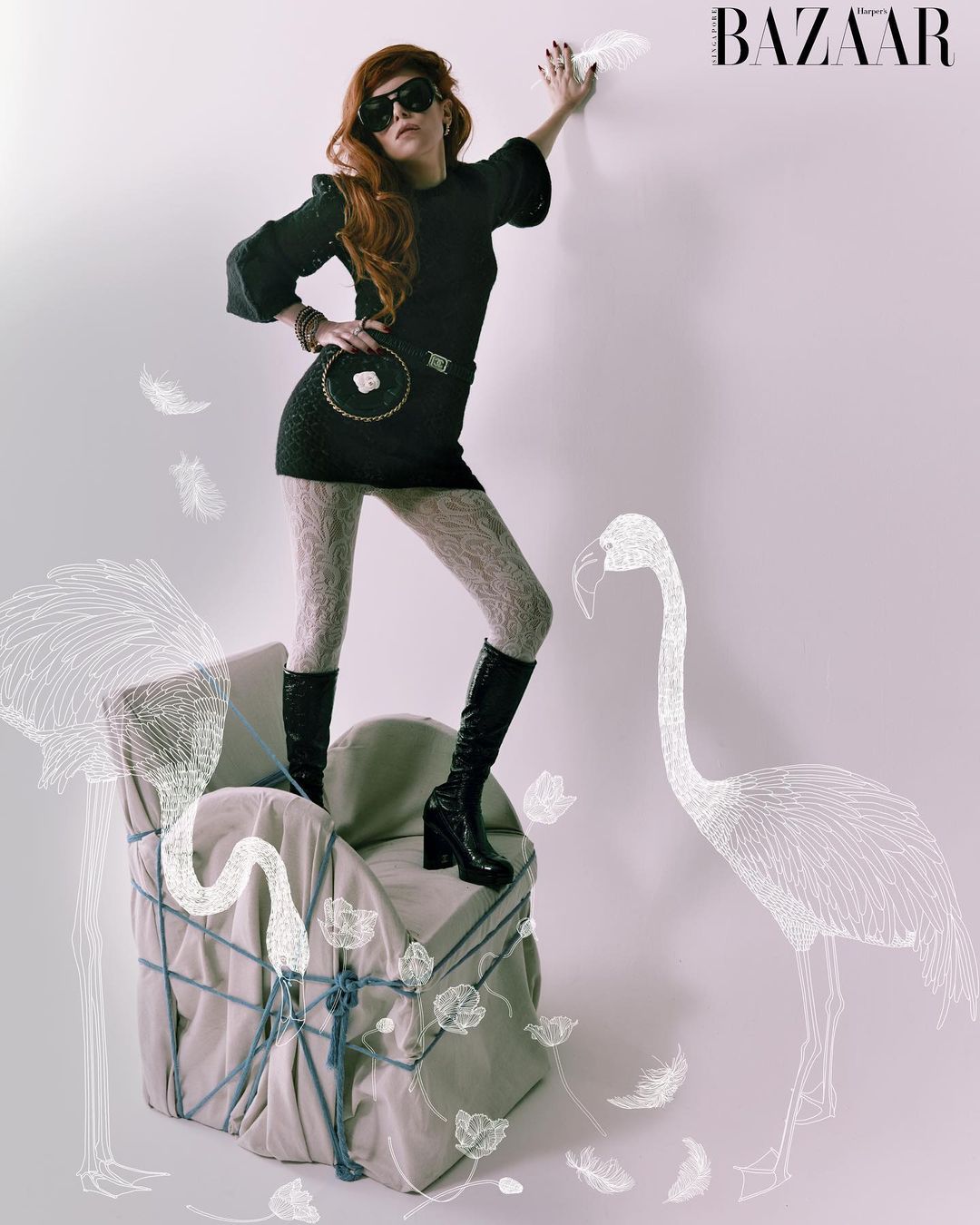 A slender redhead fashion model wearing a black mini dress, textured tights, and black platform boots strikes a pose indoors.