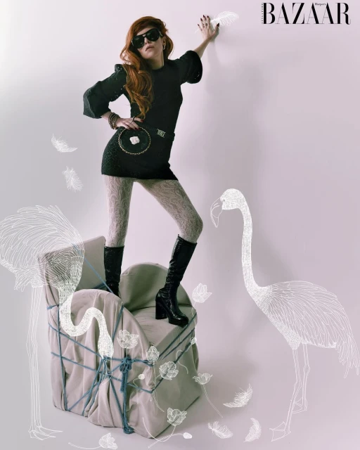 A slender redhead fashion model wearing a black mini dress, textured tights, and black platform boots strikes a pose indoors.